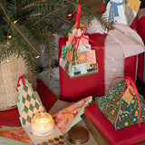 Handmade Christmas-themed gift boxes and ornaments hanging on a decorated Christmas tree and wrapped presents. The boxes feature festive patterns including checkered, tree, and holiday village designs, with one box opened to display a lit candle inside.