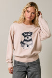 A woman wearing a light beige crewneck sweatshirt with a black and white Schnauzer dog graphic on the front, paired with gray jeans, against a solid light gray background.