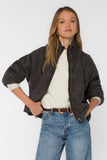 A woman modeling a black quilted zip-up jacket over a cream knit sweater and blue jeans, photographed against a plain light gray studio background.
