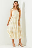 A woman modeling a sleeveless tiered midi dress with a light cream and pale yellow stripe pattern, paired with beige strappy high-heeled sandals, standing against a white wall.
