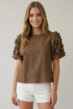 A woman wearing a brown textured short-sleeve top with dramatic ruffled puff sleeves and white denim shorts, standing against a light gray studio background.